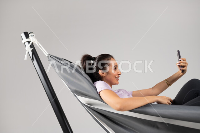 مشاركة الأخبار و الأحاديث الجيدة , إجراء مكالمة فيديو مع الأهل و الأصدقاء , بورتريه مقرب لامرأة عربية خليجية سعودية ترتدي الزي الكاجوال تستلقي على أرجوحة قماشية معلقة تمسك الهاتف المحمول بيدها , استخدام جهاز الجوال للتواصل , إيماءات الفرح و السرور , خلفية رمادية