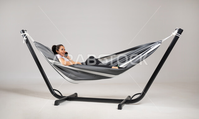 إجراء مكالمة هاتفية عبر الجوال , بورتريه لامرأة عربية خليجية سعودية ترتدي الزي الكاجوال تستلقي على أرجوحة قماشية معلقة تمسك الهاتف المحمول بيدها , مشاركة الأخبار الجيدة مع الأهل و الأصدقاء, خلفية رمادية