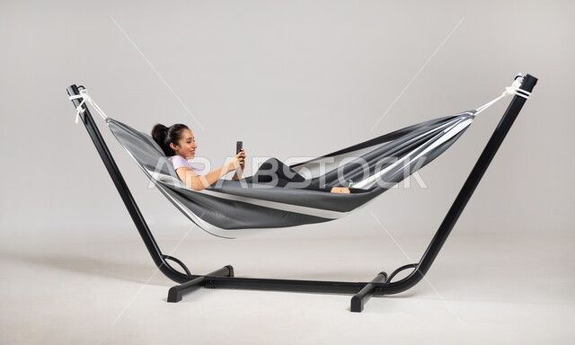 استخدام الهاتف المحمول في التسلية و الترفيه , تصفح مواقع التواصل الاجتماعي , بورتريه لامرأة عربية خليجية سعودية ترتدي الزي الكاجوال تستلقي على أرجوحة قماشية معلقة تمسك جهاز الجوال بيدها, قضاء أوقات الفراغ باستخدام الأجهزة الحديثة , خلفية رمادية