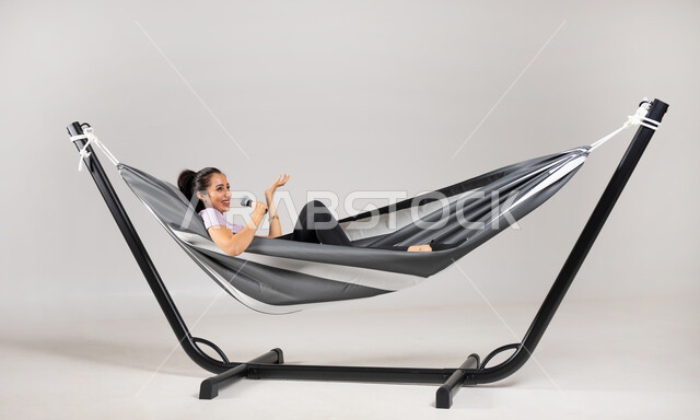 الاستمتاع بأداء العروض الصوتية , تنمية موهبة الغناء في أوقات الفراغ , بورتريه لامرأة عربية خليجية سعودية ترتدي الزي الكاجوال تستلقي على أرجوحة قماشية معلقة تمسك المايكروفون بيدها , أنشطة للتسلية و الترفيه , خلفية رمادية