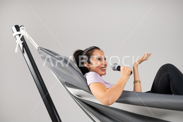 تنمية موهبة الغناء في أوقات الفراغ , بورتريه مقرب لامرأة عربية خليجية سعودية ترتدي الزي الكاجوال تستلقي على أرجوحة قماشية معلقة تمسك المايكروفون بيدها , الاستمتاع بأداء العروض الصوتية ,خلفية رمادية