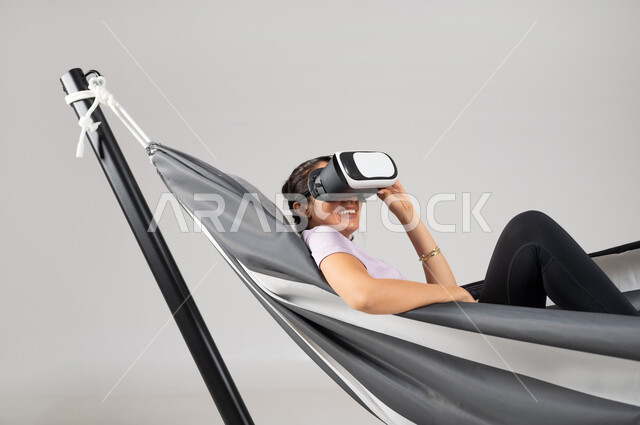 Gestures of interaction and integration, simulation of the imaginary world, close-up portrait of a Saudi Gulf Arab woman wearing casual clothing, lying on a hanging cloth hammock wearing virtual reality glasses, using advanced technologies in amusement and entertainment, gray background