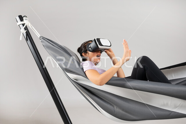 Using advanced technologies in amusement and entertainment, simulating the imaginary world, a close-up portrait of a Saudi Gulf Arab woman wearing a casual outfit, lying on a hanging cloth hammock wearing virtual reality glasses, gestures of interaction and integration, gray background