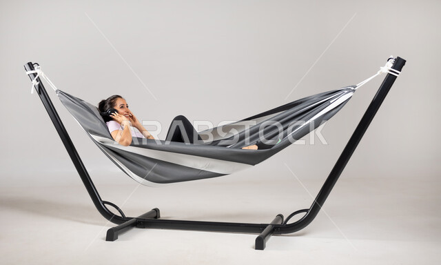 قضاء وقت الاستراحة في سماع الموسيقى المفضلة  , استخدام التقنيات الحديثة في التسلية و الترفيه , بورتريه لامرأة عربية خليجية سعودية ترتدي الزي الكاجوال تستلقي على أرجوحة قماشية معلقة تضع سماعات الرأس الصوتية , إيماءات الاستمتاع و الإصغاء ,خلفية رمادية