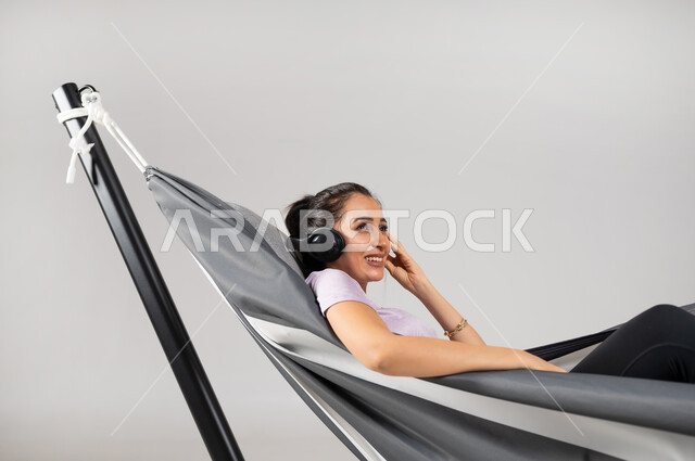 استخدام التقنيات الحديثة في التسلية و الترفيه , بورتريه مقرب لامرأة عربية خليجية سعودية ترتدي الزي الكاجوال تستلقي على أرجوحة قماشية معلقة تضع سماعات الرأس الصوتية , قضاء وقت الاستراحة في سماع الموسيقى المفضلة , خلفية رمادية