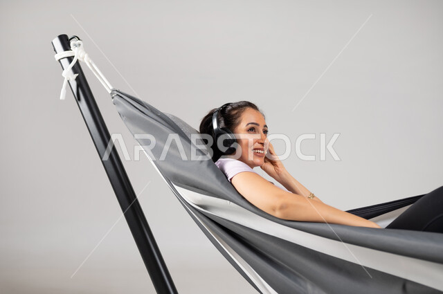 قضاء أوقات الفراغ بالاستماع إلى الأغاني , استخدام تقنية الهيدفون , بورتريه مقرب لامرأة عربية خليجية سعودية ترتدي الزي الكاجوال تستلقي على أرجوحة قماشية معلقة تضع سماعات الرأس الصوتية  , إيماءات الاستمتاع و الإصغاء , خلفية رمادية