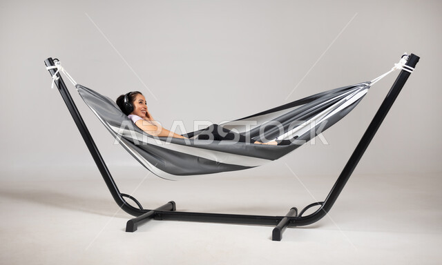 استخدام تقنية الهيدفون , بورتريه لامرأة عربية خليجية سعودية ترتدي الزي الكاجوال تستلقي على أرجوحة قماشية معلقة تضع سماعات الرأس الصوتية , قضاء أوقات الفراغ بالاستماع إلى الأغاني , إيماءات الاستمتاع و الإصغاء , خلفية رمادية