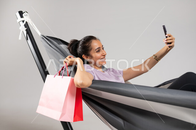 شراء الأغراض أونلاين عبر تطبيقات و مواقع على الهاتف المحمول , بورتريه مقرب لامرأة عربية خليجية سعودية ترتدي الزي الكاجوال تستلقي على أرجوحة قماشية معلقة تحمل كيس مشتريات و جوال بيديها , القيام بالتسوق الالكتروني, خلفية رمادية