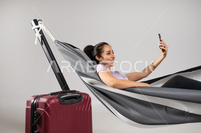 التواصل مع الأصدقاء عبر مكالمة الفيديو الهاتفية ,استخدام التقنيات الحديثة , بورتريه مقرب لامرأة عربية خليجية سعودية ترتدي الزي الكاجوال تستلقي على أرجوحة قماشية معلقة و بجانبها حقيبة سفر حمراء , التخطيط للاستمتاع بالعطلة , القيام بحجز رحلات السفر , خلفية رمادية