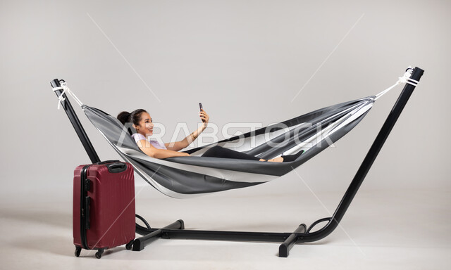استخدام التقنيات الحديثة , التواصل مع الأصدقاء عبر مكالمة الفيديو الهاتفية  , بورتريه لامرأة عربية خليجية سعودية ترتدي الزي الكاجوال تستلقي على أرجوحة قماشية معلقة و بجانبها حقيبة سفر حمراء , التخطيط للاستمتاع بالعطلة , القيام بحجز رحلات السفر , خلفية رمادية