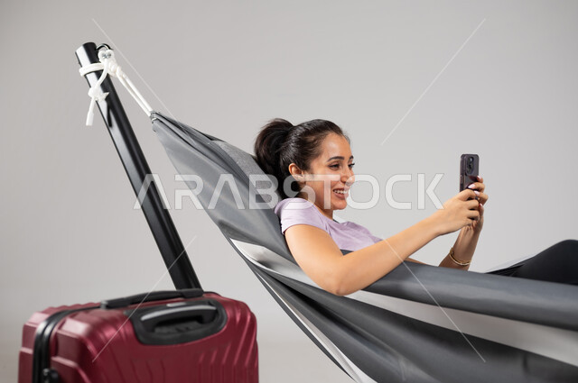 استخدام التقنيات الحديثة لحجز رحلات السفر , بورتريه مقرب لامرأة عربية خليجية سعودية تقوم بالاستلقاء على أرجوحة قماشية معلقة , التخطيط للاستمتاع بالعطلة , البحث عن أنشطة للإجازة , إيماءات الفرح و السرور , خلفية رمادية