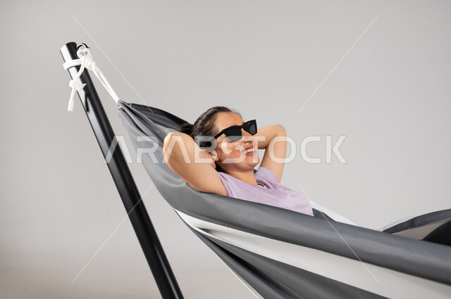 أخذ قسط من الراحة , قضاء أوقات ممتعة بالاستراحة , بورتريه مقرب لامرأة عربية خليجية سعودية ترتدي نظارات شمسية تقوم بالاستلقاء على أرجوحة قماشية معلقة  , إيماءات السعادة و السرور , خلفية رمادية