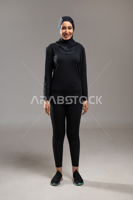 الوقوف باستقامة و استعداد , إيماءات السعادة و الاعتزاز بالنفس , بورتريه لامرأة محجبة عربية خليجية سعودية ترتدي ملابس سوداء تقف أمام الكاميرا لأخذ صورة , صورة لكامل الجسم  , خلفية رمادية