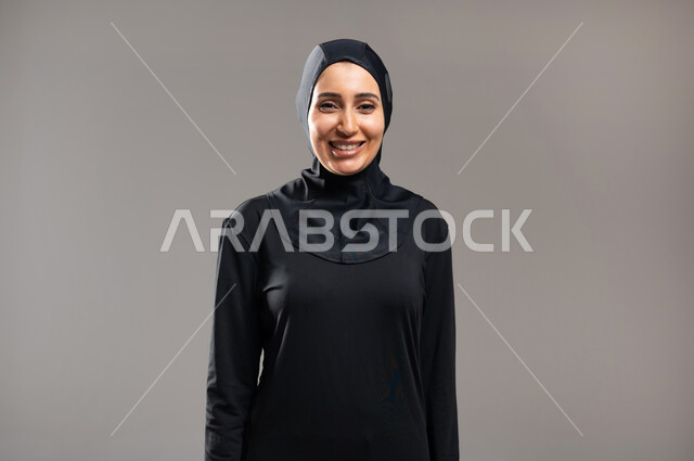 إيماءات السعادة و الاعتزاز بالنفس , الوقوف باستقامة و استعداد  , بورتريه مقرب لامرأة محجبة عربية خليجية سعودية ترتدي ملابس سوداء تقف أمام الكاميرا لأخذ صورة , خلفية رمادية