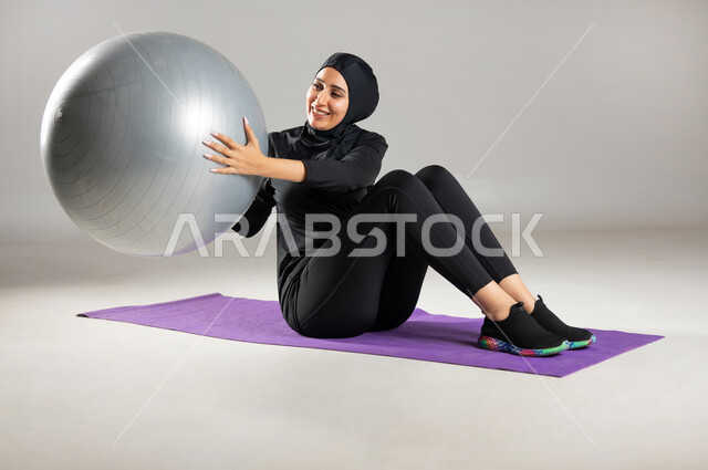 إيماءات السعادة و النشاط , مفهوم الرشاقة و اللياقة , استخدام كرة البيلاتس لممارسة التمارين الرياضية , بورتريه لامرأة محجبة عربية خليجية سعودية ترتدي ملابس رياضية , المحافظة على التوازن الجسدي  , صورة للجسم بالكامل ,خلفية رمادية