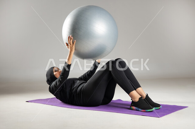 المحافظة على التوازن الجسدي , استخدام كرة البيلاتس لممارسة التمارين الرياضية , بورتريه لامرأة محجبة عربية خليجية سعودية ترتدي ملابس رياضية , مفهوم الرشاقة و اللياقة , إيماءات السعادة و النشاط , صورة للجسم بالكامل  , خلفية رمادية