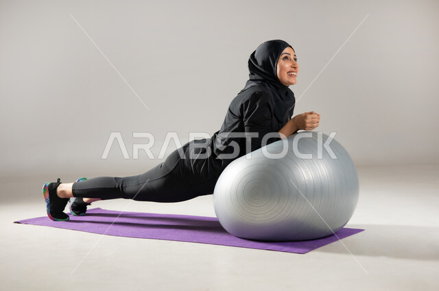 استخدام كرة البيلاتس لممارسة التمارين الرياضية , بورتريه لامرأة محجبة عربية خليجية سعودية ترتدي ملابس رياضية , المحافظة على التوازن الجسدي , مفهوم الرشاقة و اللياقة , إيماءات السعادة و النشاط , صورة لكامل الجسم  , خلفية رمادية
