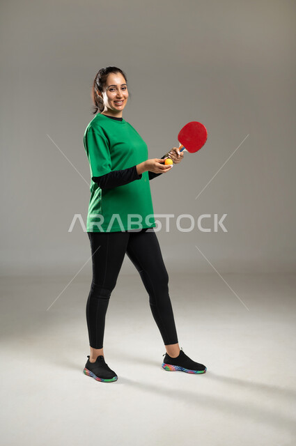 التدرب على إتقان لعبة تنس الطاولة , بورتريه لامرأة عربية خليجية سعودية ترتدي ملابس رياضية تمسك المضرب و الكرة بيديها , النظر إلى الكاميرا بإيماءات الفرح و السرور , الشغف بالألعاب الرياضية الممتعة , الفريق السعودي النسائي , صورة للجسم بالكامل , خلفية رمادية