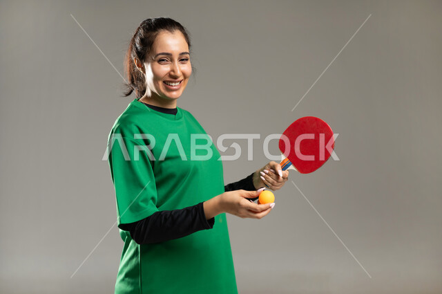 إتقان لعبة تنس الطاولة , بورتريه مقرب لامرأة عربية خليجية سعودية ترتدي ملابس رياضية تمسك المضرب و الكرة بيديها , النظر إلى الكاميرا بإيماءات الفرح و السرور , الشغف بالألعاب الرياضية الممتعة , الفريق السعودي النسائي , خلفية رمادية