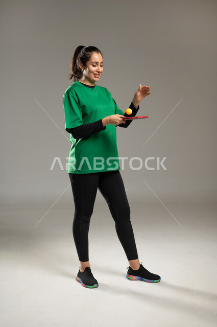 التدرب على إصابة الأهداف , ممارسة لعبة تنس الطاولة , بورتريه لامرأة عربية خليجية سعودية ترتدي ملابس رياضية تمسك المضرب و الكرة بيديها , الشغف بالألعاب الرياضية الممتعة , الفريق السعودي النسائي , صورة للجسم بالكامل , خلفية رمادية