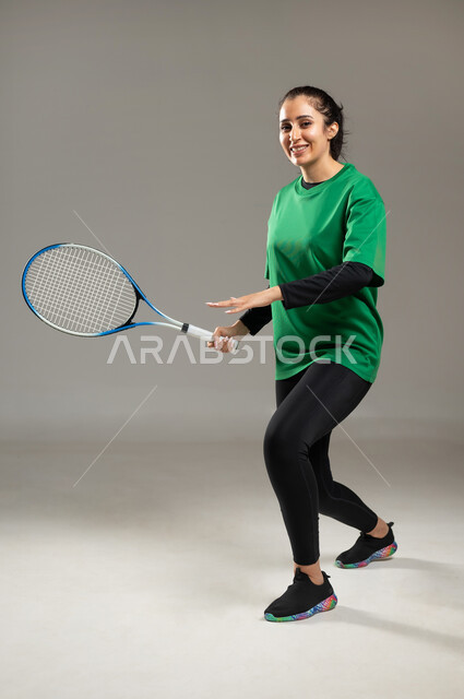قضاء أوقات الفراغ في تنمية هواية لعبة التنس , بورتريه لامرأة عربية خليجية سعودية ترتدي ملابس رياضية تمسك المضرب بيدها , النظر إلى الكاميرا بإيماءات السعادة و السرور , المحافظة على اللياقة الجسدية , صورة للجسم بالكامل , خلفية رمادية