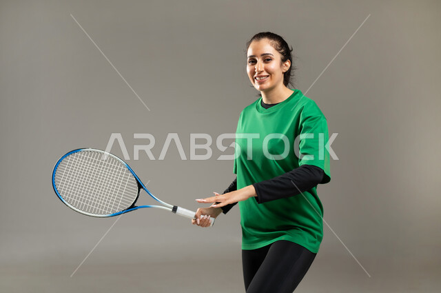 الاستمتاع بممارسة الأنشطة الرياضية , بورتريه مقرب لامرأة عربية خليجية سعودية ترتدي ملابس رياضية تمسك المضرب بيديها بإيماءات السعادة و السرور , المحافظة على اللياقة البدنية , إتقان لعبة التنس , الفريق السعودي النسائي, خلفية رمادية
