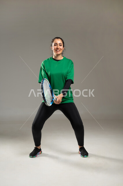 التدرب على إصابة الأهداف , القيام بالتدريبات الخاصة بلعبة التنس ,بورتريه لامرأة عربية خليجية سعودية ترتدي ملابس رياضية تمسك مضرب التنس بيديها, الفريق السعودي النسائي , صورة للجسم بالكامل , خلفية رمادية