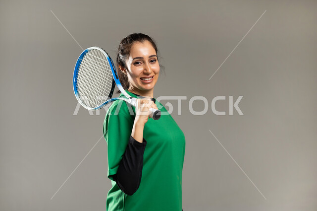 ممارسة هواية لعبة التنس , النظر إلى الكاميرا بإيماءات السعادة و الحماس ,بورتريه مقرب لامرأة عربية خليجية سعودية ترتدي ملابس رياضية , القيام بالأنشطة البدنية المسلية , إسلوب الحياة الصحي , خلفية رمادية