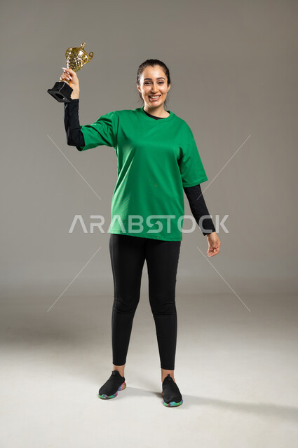النظر إلى الكاميرا بإيماءات السعادة و السرور بالفوز, الفريق السعودي النسائي ,بورتريه لامرأة عربية خليجية سعودية ترتدي ملابس رياضية تمسك الكأس بيدها تعبيرا عن الانتصار , أنشطة رياضية ترفيهية , صورة للجسم بالكامل, خلفية رمادية