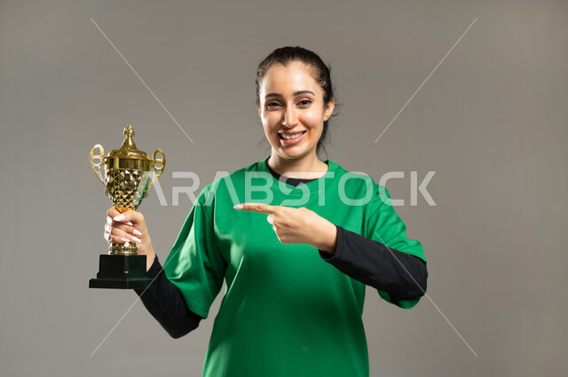 الفريق السعودي النسائي , الاستمتاع بممارسة الأنشطة الرياضية , بورتريه مقرب لامرأة عربية خليجية سعودية ترتدي ملابس رياضية , الإشارة بإصبع السبابة باتجاه الكأس بإيماءات السعادة و النصر  , خلفية رمادية