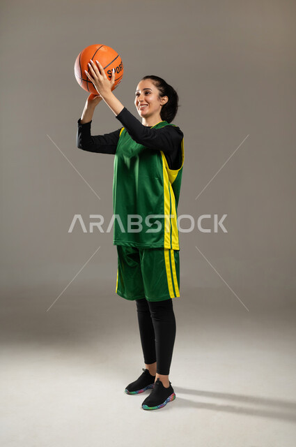 Mastering the game of basketball, women’s physical activities for fun and entertainment, portrait of a Saudi Gulf Arab woman wearing sports clothing, holding a ball in her hands, practicing hitting goals, Saudi women’s team, full-body portrait, gray background