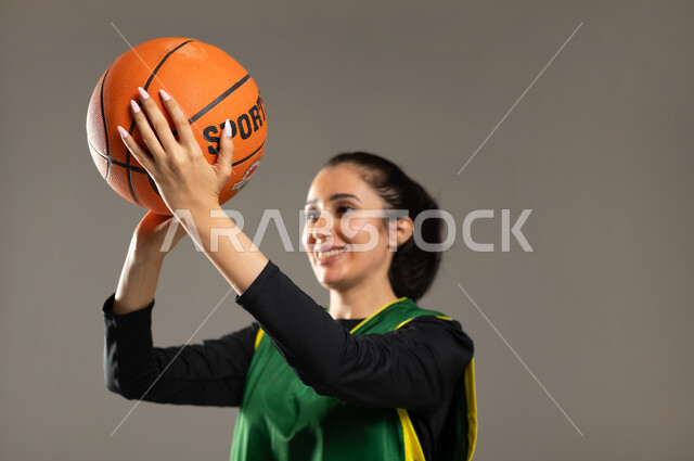 التدريب على إصابة الأهداف , إتقان لعبة كرة السلة ,الفريق السعودي النسائي , بورتريه مقرب لامرأة عربية خليجية سعودية ترتدي ملابس رياضية تمسك الكرة بيديها بإيماءات الفرح و السعادة , تعلم , خلفية رمادية