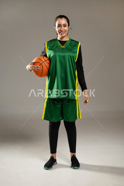 ممارسة هواية لعبة كرة السلة , أنشطة رياضية للتسلية و الترفيه , بورتريه لامرأة عربية خليجية سعودية ترتدي ملابس رياضية تقف باستعداد تمسك الكرة بيدها مع إيماءات السعادة و السرور , الفريق السعودي النسائي , صورة للجسم بالكامل , خلفية رمادية