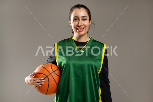 Women's sports activities in Saudi Arabia, practicing the hobby of basketball, a close-up portrait of a Saudi Gulf Arab woman wearing sports clothing holding the ball with gestures of happiness and pleasure, the Saudi women's team, gray background