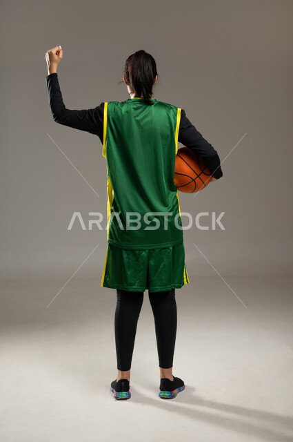 الشغف بالألعاب الرياضية , ممارسة هواية كرة السلة , بورتريه من الخلف لامرأة عربية خليجية سعودية ترتدي ملابس رياضية تمسك الكرة بإيماءات التشجيع و الحماس , الفريق السعودي النسائي , صورة للجسم بالكامل , خلفية رمادية