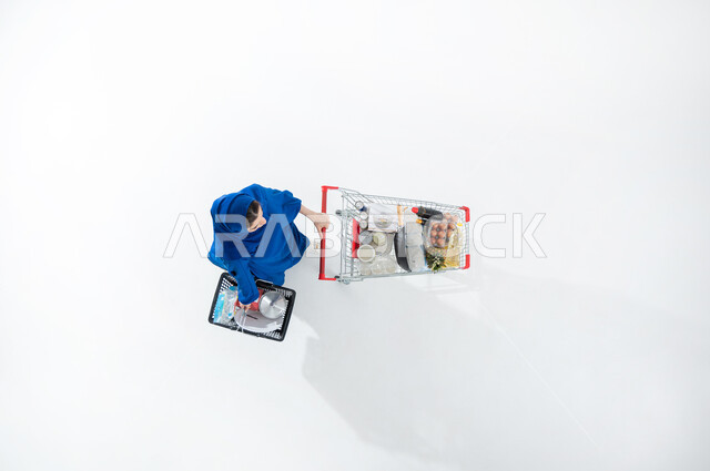 قضاء الاوقات الممتعه في المراكز التجارية بالمملكة العربية السعودية، شراء الاحتياجات الضروريه المنزليه، بورتريه عامودي علوي لسيده عربية خليجية سعودية محجبه ترتدي العباءة الملونه العصريه تقوم بجرعربة التسوق وحمل سله التسوق الغذائيه، خلفيه بيضاء