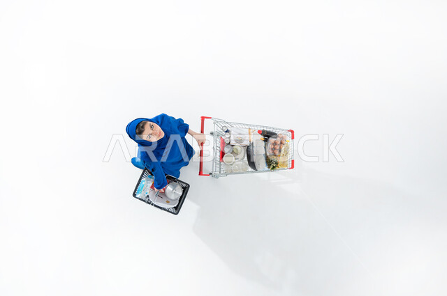 قضاء الاوقات الممتعه في المراكز التجارية بالمملكة العربية السعودية، شراء الاحتياجات الضروريه المنزليه، بورتريه عامودي علوي لسيده عربية خليجية سعودية محجبه ترتدي العباءة الملونه العصريه تقوم بجرعربة التسوق وحمل سله التسوق الغذائيه، خلفيه بيضاء