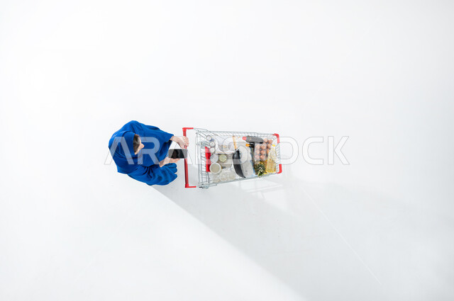 قراءه قائمه المستلزمات بواسطه الهاتف المحمول، شراء اللوازم والاحتياجات اليوميه، بورتريه عامودي علوي لسيدة عربية خليجية سعودية محجبه ترتدي العباءة الملونه تجر بيدها عربه التسوق الممتلئه بالاغذيه تحمل بيدها هاتفها المحمول، الاستفاده من العروض والتخفيضات، خلفيه بيضاء