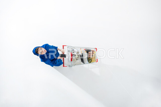 النظر إلى شيء ما والاستعداد للتسوق وشراء الحاجات من المراكز التجارية، الاستفادة من التخفيضات والعروض على المنتجات، بورتريه عامودي علوي لسيدة عربية خليجية سعودية محجبه ترتدي العباءة الملونه العصريه تقوم بجرعربه التسوق تحمل بيدها هاتفها المحمول، قضاء وقت ممتع في التسوق، خلفيه بيضاء