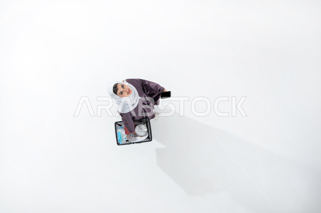 البحث عن المستلزمات في المراكز التجارية السعوديه، التسوق وشراء الاحتياجات المنزليه الضروريه، بورتريه عامودي علوي لسيدة عربية خليجية سعودية ترتدي العباءة الرمادية والحجاب تمسك بيدها سلة التسوق تشير بجهازها المحمول بشاشه سوداء، خلفيه بيضاء