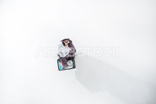 Searching for supplies in Saudi malls, taking advantage of discounts and reductions on products, vertical portrait of a Saudi Arabian Gulf woman wearing a gray abaya and hijab holding a shopping basket in her hand, talking to someone on her mobile device, white background