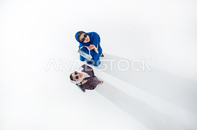مواكبة تصاميم الأزياء والموديلات الحديثة, بورتريه عامودي علوي لشابتان عربيتان خليجيتان محجبتان ترتديان العباءات الملونه العصريه والنظارات الشمسيه تحملان حقائب شخصيه ملونه بايماءات الأنوثة واللطافة, التسويق للمنتجات السعودية والماركات العالمية الفاخرة, خلفيه بيضاء