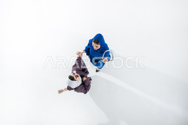 قضاء أوقات ممتعة مع الأصدقاء، بورتريه عامودي علوي لصديقتان عربيتان خليجيتان سعوديتان محجبتان  ترتديان العباءه العصرية الملونه والنظارات الشمسية تشيران بايماءات الفرح والسرور، الاشاره الى شئ ما، أحدث الأزياء النسائية المعاصرة، مفهوم الموضة والأناقة، خلفية بيضاء