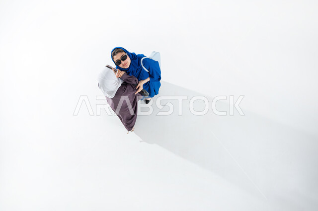 النميمة والتحدث عن شخص ما، ايماءات الانصات والتركيز بالكلام، التحدث بصوت منخفض، بورتريه مقرب لشابة عربية خليجية سعودية محجبه  ترتدي النظاره الشمسيه والعباءة العصريه الملونه تهمس بإذن صديقتها، مفهوم الكتمان وحفظ الأسرار بين الزملاء، خلفيه بيضاء