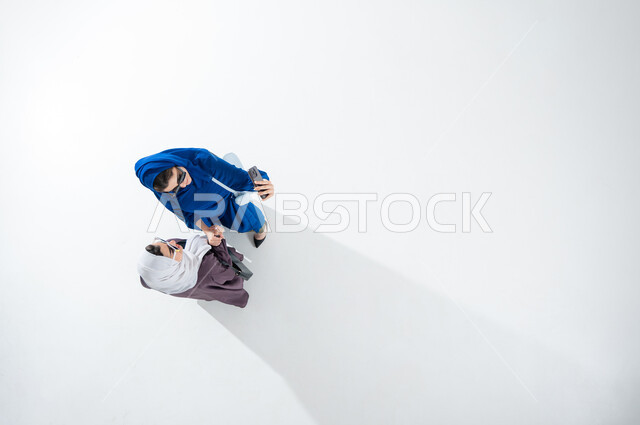 توثيق اللحظات السعيدة، استخدام الهاتف المحمول والتقنيات الحديثه، بورتريه عامودي علوي لشابة عربية خليجية سعودية محجبة ترتدي العباءة العصريه الملونه والنظاره الشمسيه تقوم بالتقاط صور السيلفي التذكارية مع صديقتها بإيماءات السعادة والسرور، خلفية بيضاء