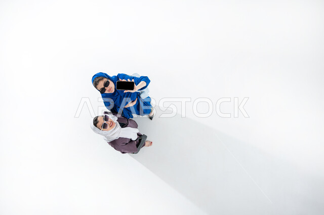 عرض التطبيقات من خلال الهاتف المحمول، بورتريه عامودي علوي لسيدة عربية خليجية سعودية تحمل بيدها الهاتف المحمول بشاشه سوداء ، شابة خليجية سعودية مبتسمة تقف بجانبها ترتديان العباءه الملونه العصريه والنظارات الشمسيه، التسويق الرقمي ، اعلانات تسويقية ترويجية ، خلفية بيضاء