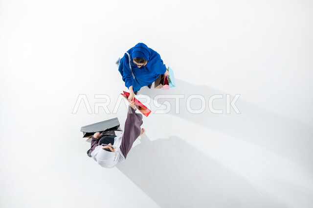 Take advantage of seasonal offers and discounts, vertical overhead portrait of two Saudi Arabian Gulf ladies wearing colorful modern abaya and sunglasses carrying colorful shopping bags, shopping and buying supplies from malls in Saudi Arabia, Black Friday offers, white background