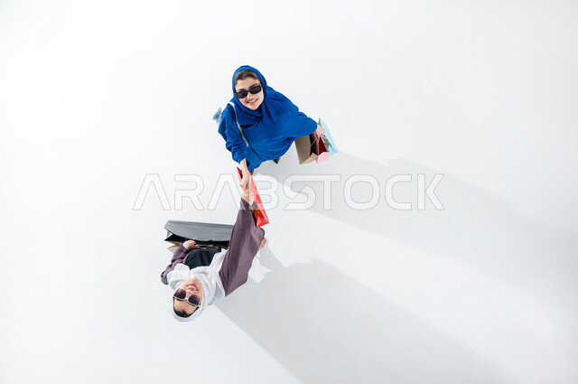 Take advantage of seasonal offers and discounts, vertical overhead portrait of two Saudi Arabian Gulf ladies wearing colorful modern abaya and sunglasses carrying colorful shopping bags, shopping and buying supplies from malls in Saudi Arabia, Black Friday offers, white background