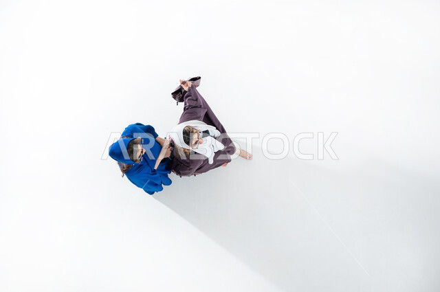 متعة الخروج مع الأصدقاء، ايماءات اللطافة والظرافة، بورتريه عامودي علوي لسيدتان عربيتان خليجيتان سعوديتان محجبتان ترتديان عباءات عصريه ملونه والنظاره الشمسيه تحملان بيدهما شنطهما الشخصيه، قضاء الوقت مع الاخوات في الخارج، ايماءات السعاده والسرور، خلفيه بيضاء