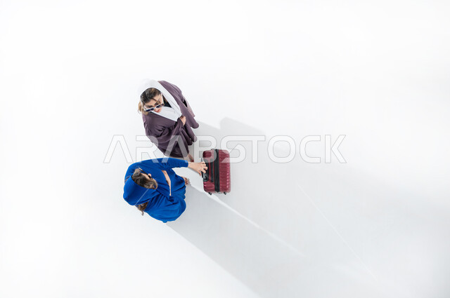 الاستمتاع بإجازة الصيف والهروب من ضغوط الحياة، الاستعداد للسفر الممتع, الاهتمام بالموضة والأزياء العصرية، بورتريه عامودي علوي لسيدتان عربيتان خليجيتان سعوديتان محجبتان ترتديان العباءه الملونه والنظارة الشمسيه تمسك حقيبة الأمتعة بإيماءات السرور والمتعة, خلفيه بيضاء